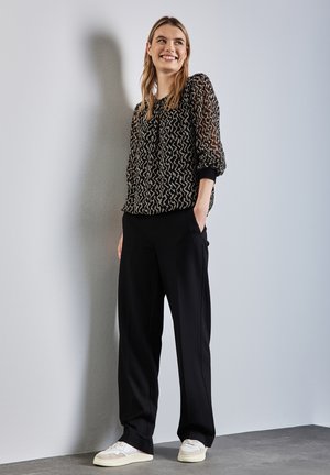 Blusa negra de manga larga transparente con un patrón geométrico, combinada con pantalones anchos negros y zapatillas crema. Texturas suaves y ajuste relajado.