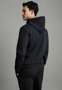 Hanorac negru cu siret, fabricat din material moale. Dispune de un buzunar tip kangaroo și manșete cu jumătate de gumă. Este purtat cu pantaloni negri asortați.