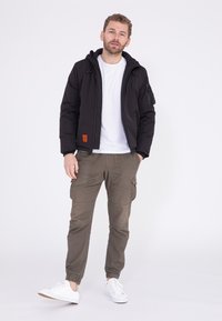 Zwarte geïsoleerde jas met capuchon, ritsluiting en oranje label; gecombineerd met olijfgroene cargobroek en witte sneakers. Strak, modern ontwerp.