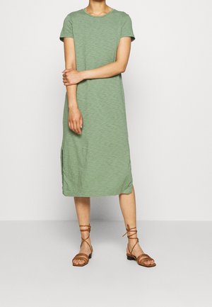 Person wearing a knee-length, short-sleeve green dress and brown strappy flat sandals, standing with one arm crossed over the other.