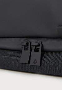 Fekete nejlon hátizsák közeli felvétel, két cipzárral, amelyek téglalap alakú húzókkal rendelkeznek, texturált anyagból készült, és diszkrét márkázással a cipzár húzón.