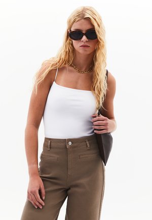 Top bianco con spalline sottili, pantaloni verde oliva a vita alta con tasche frontali, collana a catena dorata e occhiali da sole con lenti nere.