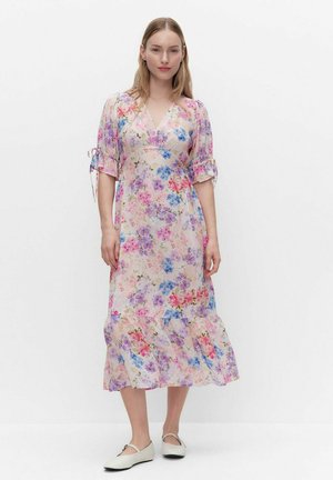 Kvinde iført en pastelblomstret midikjole med puffede halværmer bundet ved albuerne og hvide flade Mary Jane-sko.