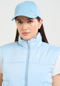 Šviesiai mėlyna pūkinė liemenė su aukštu apykakle ir priekiu užtraukiama, derinama su atitinkama šviesiai mėlyna kepure, turinčia logotipą. Lygi tekstūra, modernus dizainas.