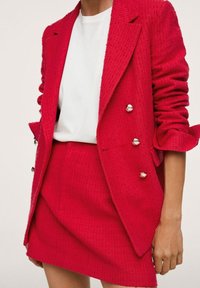 Blazer rojo texturizado con un diseño de doble botonadura y botones metálicos, combinado con una minifalda a juego. Camiseta blanca debajo.