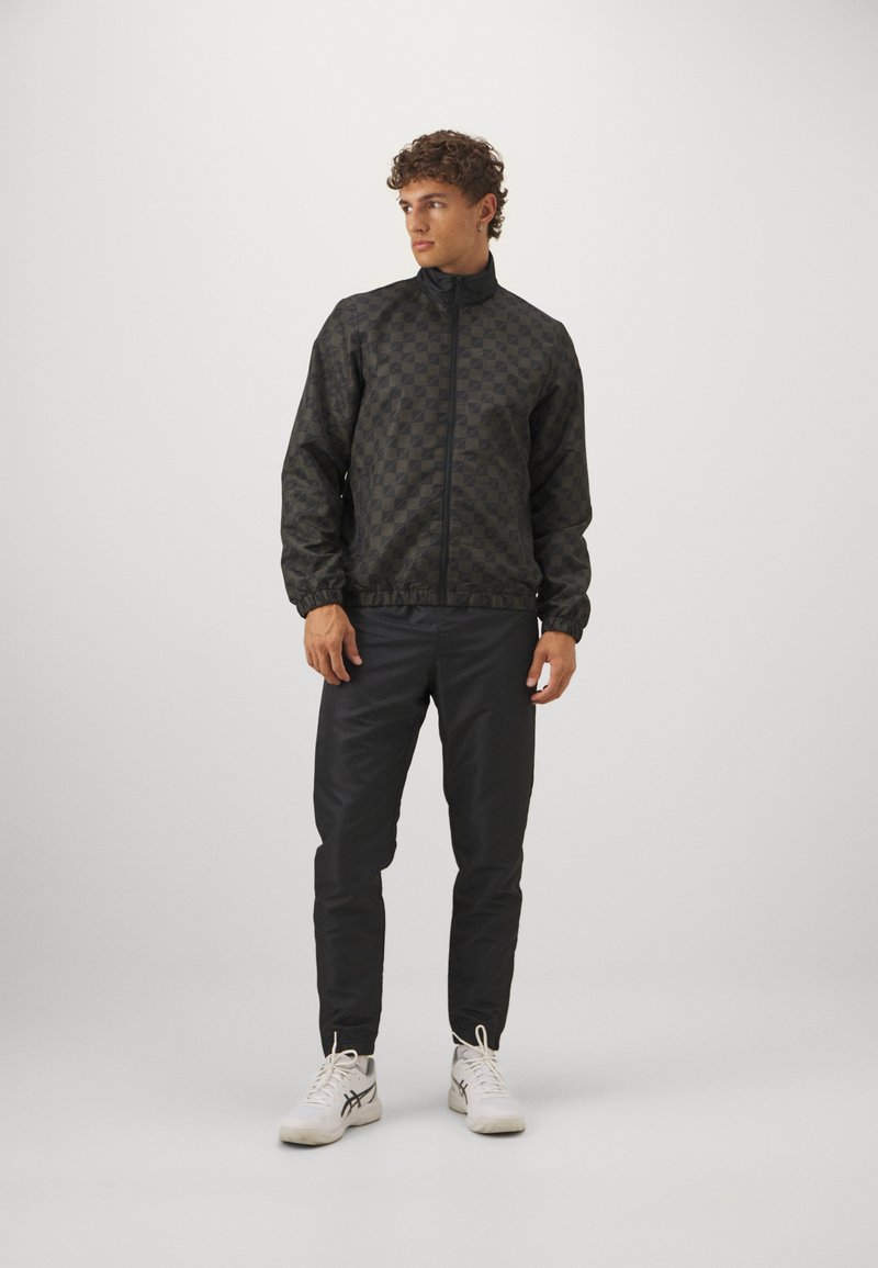 Giacca nera con motivo, colletto alto, polsini ed elastico in vita. Indossata con pantaloni scuri e scarpe da ginnastica bianche.