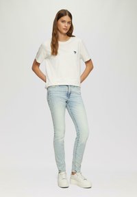 Weißes T-Shirt mit kurzen Ärmeln und einem kleinen blauen Herz-Logo, kombiniert mit hellblauen Skinny-Jeans und weißen Plateausneakern.