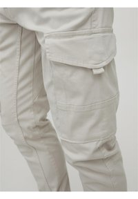 Beige cargo-housut, joissa on läppätasku reidessä ja yksityiskohtaiset tikkaukset saumoissa ja taskuissa.