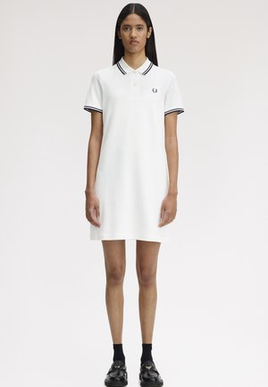 Mujer joven de pie, vestida con un vestido polo blanco con ribete negro en el cuello y las mangas, calcetines negros y mocasines negros sobre un fondo liso.