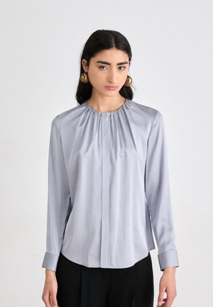 Lys grå langermet bluse i glatt stoff med rynket hals og knappelukking. Har mansjetter med en liten flare.