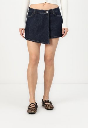 Forfra af en person iført en mørk asymmetrisk denimnederdel, hvid kort striktrøje og leopardmønstrede flade sko med stropper.