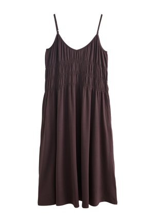 Vrouw met lang donker haar, gekleed in een donker mouwloze jurk en schelpenketting, staat op een rotsachtige oever met oceaan op de achtergrond.