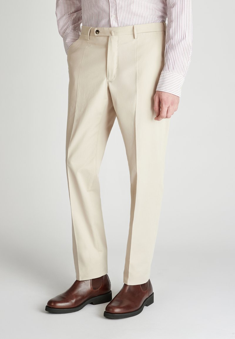 Beige Stoffhosen mit geradem Schnitt, ausgestattet mit Seitentaschen, einem Reißverschluss und einer Stofftextur. Kombiniert mit braunen Leder-Stiefeln bis zum Knöchel.