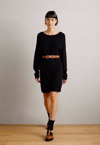 Robe noire en tricot à manches longues, dotée d'une ceinture marron avec une boucle en argent à la taille. Modèle portant des chaussures marron avec des accents noirs.