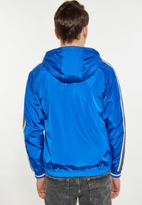 Jachetă de vânt din nylon albastru, cu glugă, manșete și tiv elastic, având dungi albe contrastante pe mâneci.