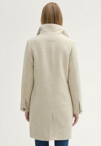 Beige gyapjúpálca, magas gallérral, oldalsó zsebekkel és gombos mandzsettákkal. A háta középen varrott és a nyakán címke található.