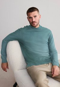 Lys grøn strikket sweater med høj krave og ribbede manchetter, parret med beige bukser. Sidder på en struktureret, lys stol.