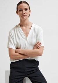 Biela blúzka s krátkym rukávom a výstrihom do V so subtílnymi vertikálnymi pruhmi, spárovaná s čiernymi oblekovými nohavicami. Hladká tkanina, voľný strih.