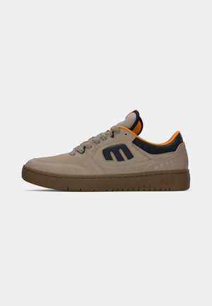 Sneaker in suede beige con accenti blu scuro, fodera arancione e suola in gomma marrone; presenta un logo sul lato e perforazioni per la traspirabilità.