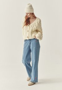Cardigan scurt din tricot cu motive cable-knit de culoare crem, cu nasturi mari, purtat cu blugi largi de culoare bleu deschis, decorati cu broderii florale colorate si adidași bej.