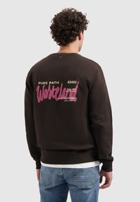 Krullenkop die een donkerbruin sweatshirt draagt met roze en beige tekst op de rug, gecombineerd met lichtblauwe jeans.