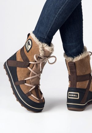 Brune og sorte vinterstøvler med pelsfor og beige snørebånd båret sammen med mørkeblå skinny jeans, vist fra siden og bagfra.