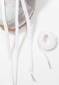 Gros plan d'une basket grise avec une section de lacets blancs non noués avec un fil doré à côté d'un lacet blanc enroulé proprement sur un fond blanc.