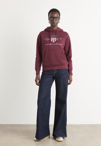 Felpa con cappuccio bordeaux con tasca frontale e logo GANT; abbinata a jeans a gamba larga blu scuro e scarpe di colore chiaro.