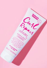 Curl Repair shampoo van Umberto Giannini in een witte tube met roze tekst. Kenmerken zijn onder andere de belofte om beschadigde krullen te herstellen, met een inhoud van 250 ml.