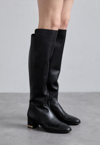 Botas negras hasta la rodilla de cuero suave, con tacón cuadrado y un sutil acento dorado en el tacón. Diseño minimalista sin patrones.