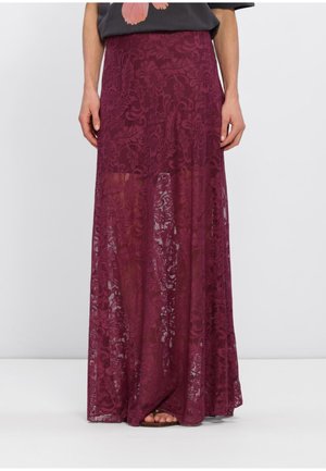 Femme portant une longue jupe en dentelle bordeaux avec des motifs floraux, associée à un haut noir et des sandales.