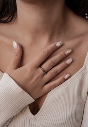 Weicher beiger Rippenpullover mit V-Ausschnitt. Hand, die auf der Brust ruht, zeigt einen dünnen Silberring und manikürte Nägel mit floralen Mustern in sanften Farben.