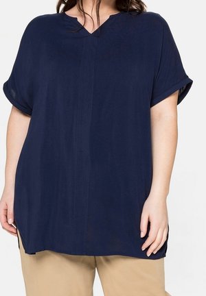 Vrouw met een loszittende marineblauwe korte mouwen blouse met een ingezette halslijn en beige broek tegen een lichte achtergrond.
