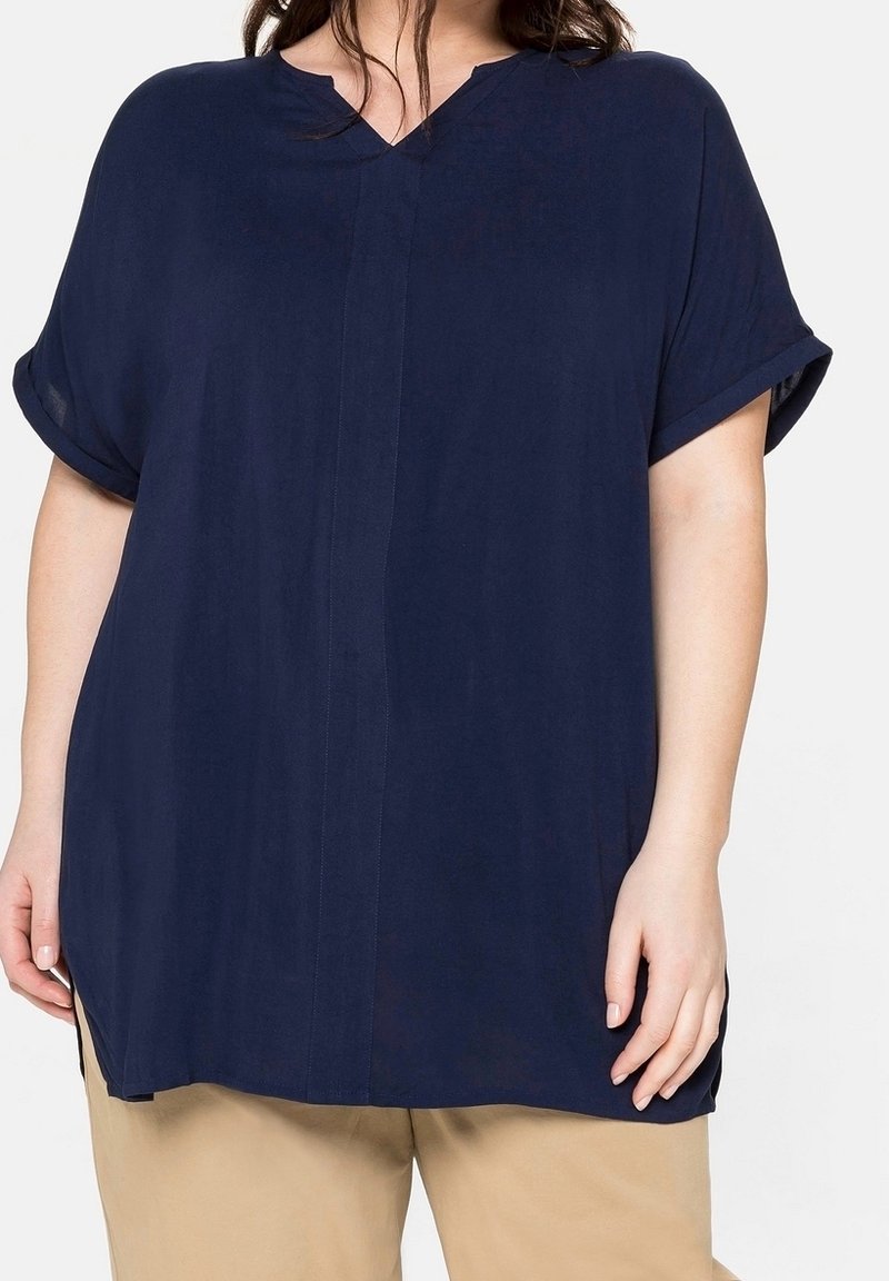 Femme portant un chemisier bleu marine ample à manches courtes avec un col cranté et un pantalon beige sur un fond clair.