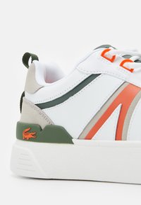 Vit sneaker med orange och gröna accenter, grön hälflik, orange varumärkeslogotyp på grön sula, samt vita snören med orange öglor.