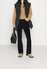 Svart pufferväst med huva, oversize beige kabelstickad tröja, svarta manchesterbyxor och beige blockklackskor. Tillbehör inkluderar en strukturerad svart väska.