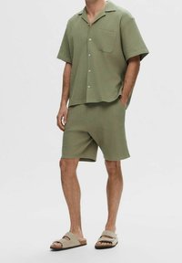 Grön rutigt kortärmad skjorta med ficka, matchad med kortbyxor. Bärs med beiga slip-in sandaler. Lättviktigt tyg och avslappnad passform.