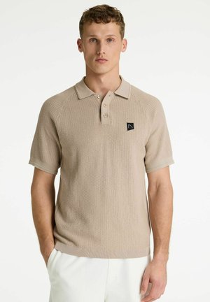 Mladý muž nosí béžové textúrované polo tričko s krátkym rukávom a gombíkmi a biele nohavice, stojaci pred jednoduchým bielym pozadím.