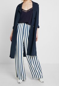 Marineblauer Mantel über einem transparenten Oberteil mit Spitzenkragen, kombiniert mit weißen und marineblauen, breit geschnittenen Hosen mit Streifen und weißen Schuhen mit Absatz.