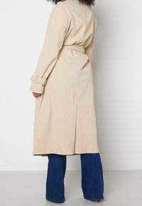 Beżowy płaszcz trenczowy z długimi rękawami, paskiem do zawiązania i szczelinami po bokach. Materiał wygląda na lekki i ma gładką teksturę. W zestawieniu z niebieskimi spodniami dzwonami.