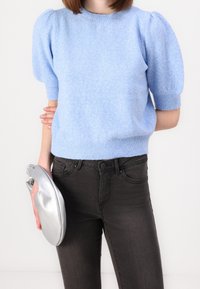 Ljusblå, texturerad stickad tröja med puffärmar och korta ärmar, kombinerad med mörka slim-fit jeans. Håller en silvermetallisk clutchväska.