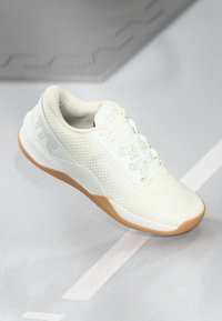 Witte sportschoen met een bovenkant van mesh en een gumrubberen zool, geplaatst op een grijze ondergrond met witte lijnen.