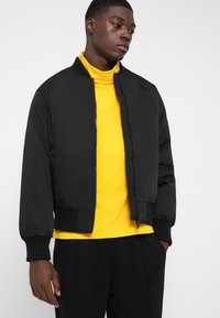 Jauns vīrietis, ģērbies melnā bomber jakā pār spilgti dzeltenu turtleneck kreklu, ar melnām biksēm, stāvot pret vienkārša balta fona.