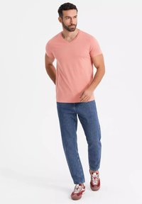 T-shirt rosa chiaro con scollo a V abbinata a jeans blu a gamba dritta e sneaker rosse. La maglietta presenta una texture morbida e una vestibilità casual.