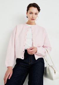 Ροζ καζάκ με απαλή υφή, με στρογγυλή λαιμόκοψη, μπροστινό κούμπωμα, ριμπ λάστιχο στα μανίκια και cropped εφαρμογή. Φοράει από πάνω ένα λευκό πουκάμισο.