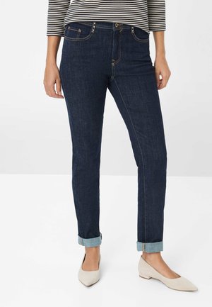 Dunkelblaue Denim-Jeans im Slim-Fit-Schnitt, mit umgeschlagenen Säumen, goldenen Nähten und vier Taschen. Dazu beigefügte beige spitze Ballerinas.