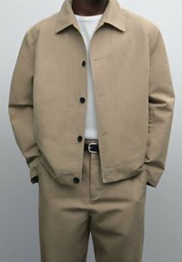 Giacca di cotone beige con colletto, chiusura a bottoni e due tasche frontali, abbinata a pantaloni coordinati e una camicia bianca sotto.