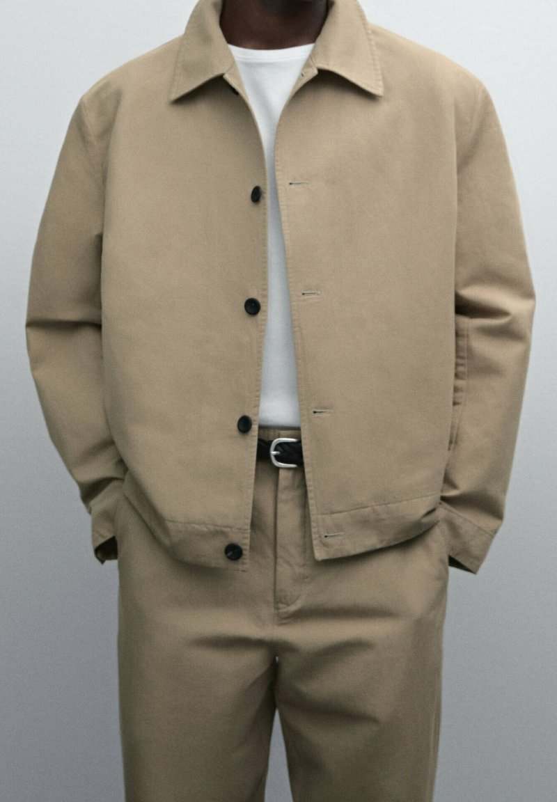 Giacca di cotone beige con colletto, chiusura a bottoni e due tasche frontali, abbinata a pantaloni coordinati e una camicia bianca sotto.