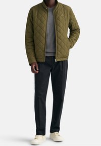 Giacca imbottita verde oliva con colletto alto e maglione grigio, abbinata a pantaloni scuri e sneakers color crema. Design semplice con accenti minimi.