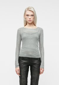 Jauna moteris su ilgais šviesiais plaukais vilkintis prigludusius pilkus ilgomis rankovėmis marškinėlius ir juodas tekstūruotas kelnes, stovinti priešais vientisą baltą foną.